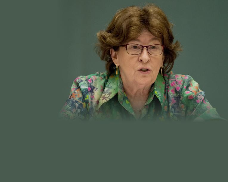 A woman with short brown hair and glasses speaks, wearing a green floral blouse against a teal background. Partially obscured.