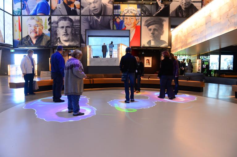 A group of people are standing within part of a white circle. Blue and purple lights are projected onto the floor surrounding each person. Partially obscured.