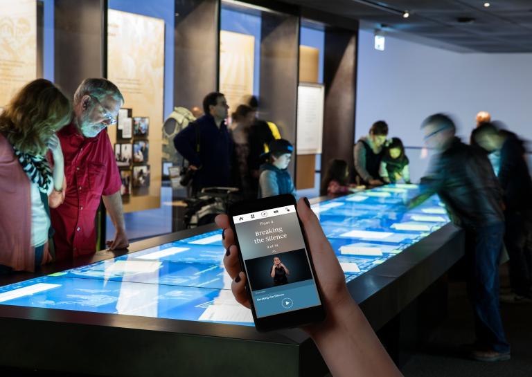 A hand holds up a mobile phone. On the phone are the words “Floor 4” and “Breaking the Silence,” and a person speaking using sign language. In the background, Museum visitors are looking at a large rectangular table with a touchscreen. Partially obscured.