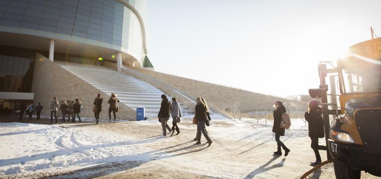 It is a bright winter day. A group of young adults are exiting a yellow school bus (right) and are walking toward the Museum entrance. Partially obscured.