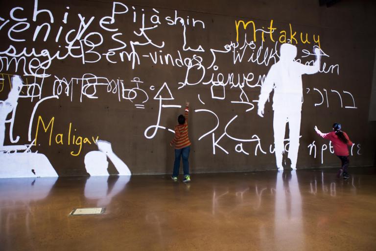 A video of people’s silhouettes is projected onto a large wall. They are writing the word “welcome” in many languages. Two children interact with the video. Partially obscured.