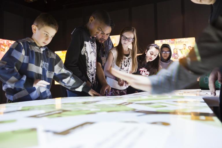 A group of students look at the top of an interactive table emitting white light. Partially obscured.