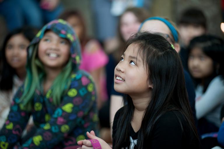 A girl lifts her head and eyes slightly upward. She has a smile on her face. Behind her are more children smiling. Partially obscured.