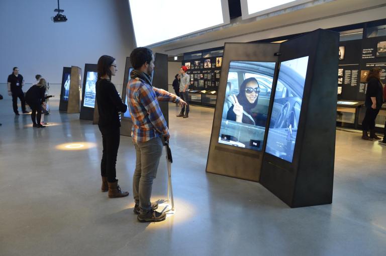 A man and woman stand looking at a double-sided video screen in a museum gallery. The man is pointing at the screen. Partially obscured.