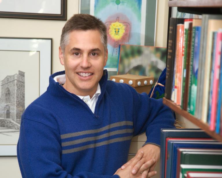 A man stands next to a bookshelf and looks into the camera. Partially obscured.