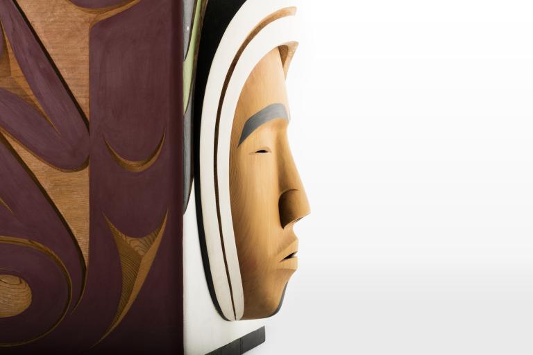 A closeup of a carved wooden box, showing the carved face of a person against a white background. Partially obscured.