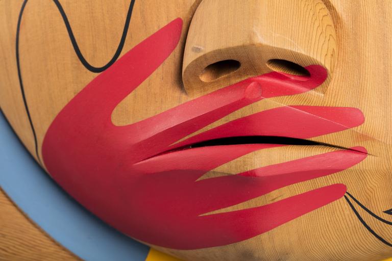 A closeup of a carved wooden box, showing a painted image of a red hand over a carved mouth. Partially obscured.