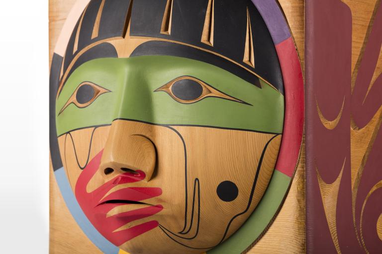 A closeup of a carved wooden box, showing the carved face of a person with a painted red hand over their mouth. Partially obscured.