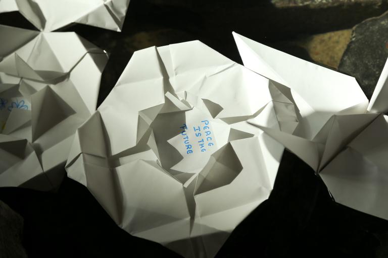  Folded paper lilies floating in water. “Peace is the future” is written in the centre of one lily. Partially obscured.