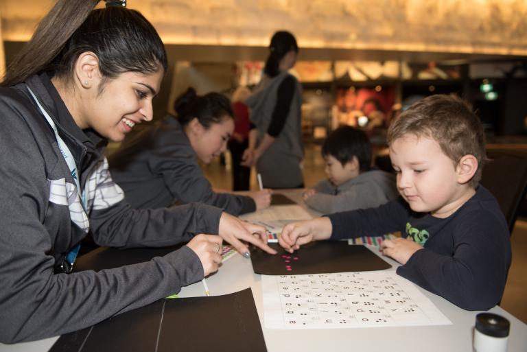 A Museum staff member helping a young child place coloured dots in the shape of braille letters. Partially obscured.