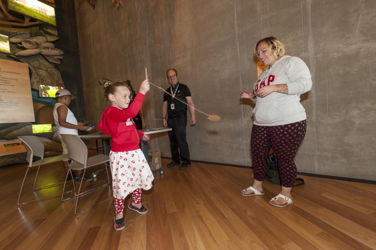 A young girl holds a small stick attached to a string and a small circular piece of cardboard. She is swinging this rope and cardboard as two adults watch. Partially obscured.