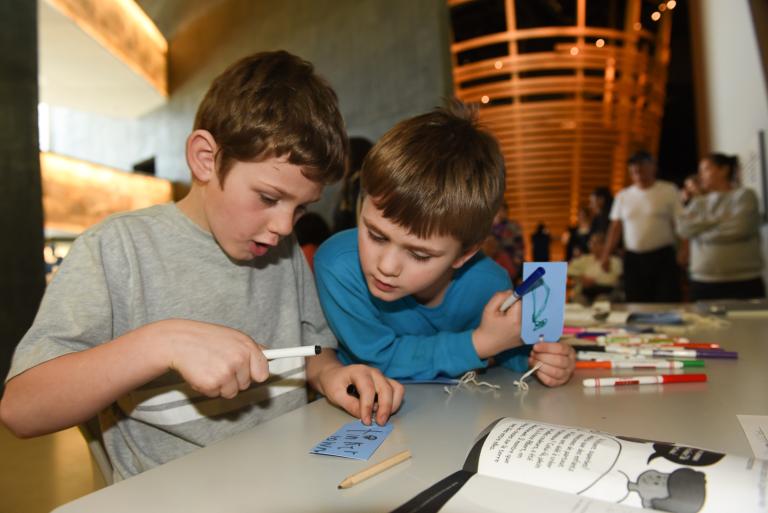 Two young boys holding markers and small cards with string and reading one of the cards. Partially obscured.