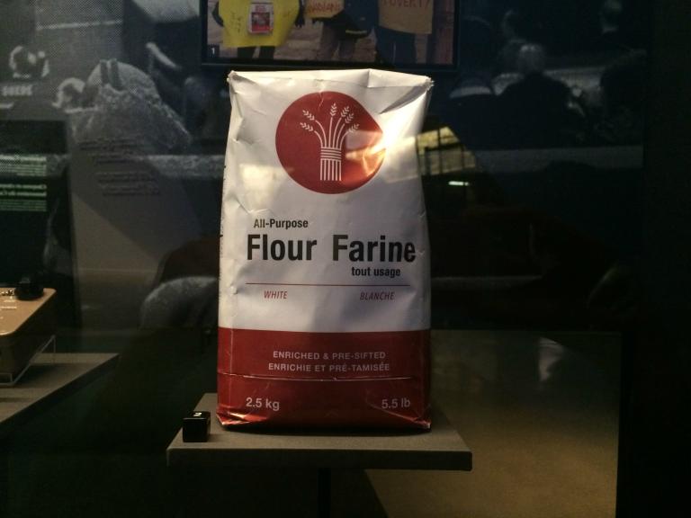A bag of flour on display. The text reads: All-Purpose Flour/Farine tout usage, White/Blanche, Enriched & Pre-Sifted/Enrichie et pré-tamisée, 2.5 kg/5.5lb