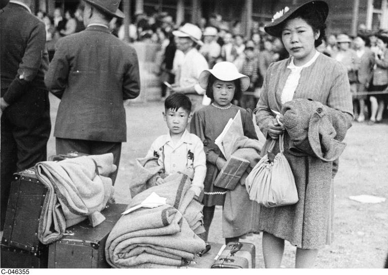 A black and white image of a woman and two children standing behind a pile of luggage and blankets and looking at the camera. Partially obscured.