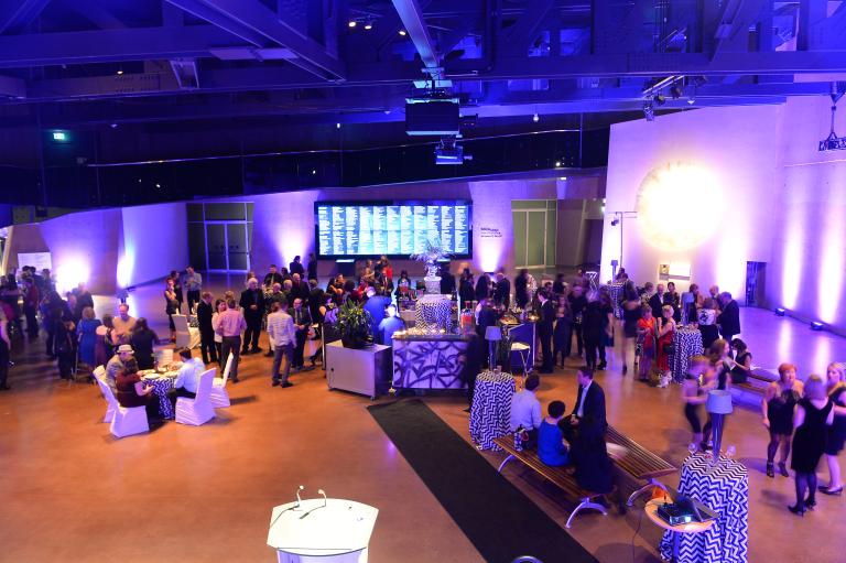 A party is taking place in a large room. Guests are gathered around tables holding glasses and chatting. Purple and blue light is cast throughout the room. Partially obscured.