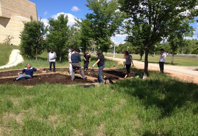 Seven men and women work in a garden on a sunny day. The garden is circular and has very few plants in it. Around the garden are tall grass, trees, a walking path and a sidewalk. In the background, part of the Museum’s stone structure can be seen. Partially obscured.