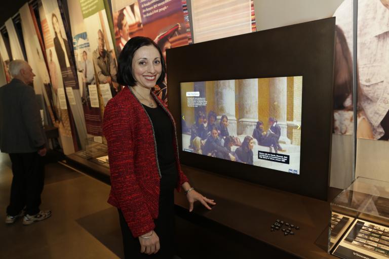 Marina Nemat smiles and stands beside a large digital display screen. She is wearing a red jacket and is looking directly at the photographer. The screen features a picture of people sitting on steps in front of large stone columns. Partially obscured.