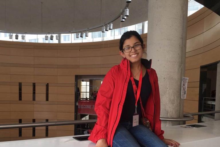 A young woman sits on a ledge in a large circular hall. She is smiling at the camera and wearing jeans, a dark blouse and a red jacket Partially obscured.