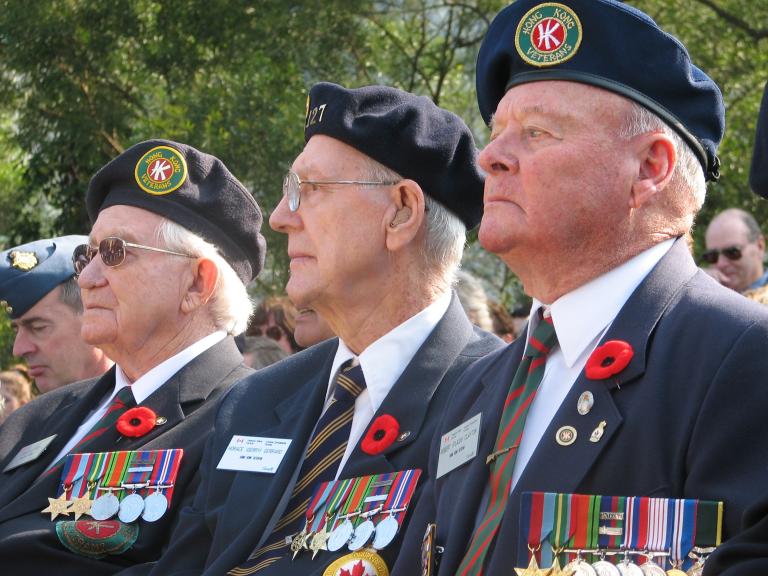 Three older men in Canadian Legion uniforms sit beside each other. They are all wearing Remembrance Day poppies on their uniforms. Partially obscured.
