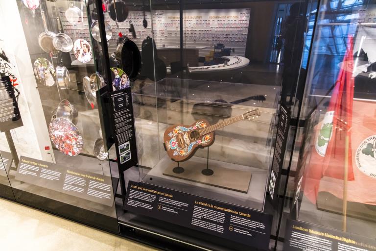 A row of museum display cases with various artifacts inside. A guitar is displayed under glass. The guitar has a colourful floral print with a bird hand-painted onto its front. Partially obscured.