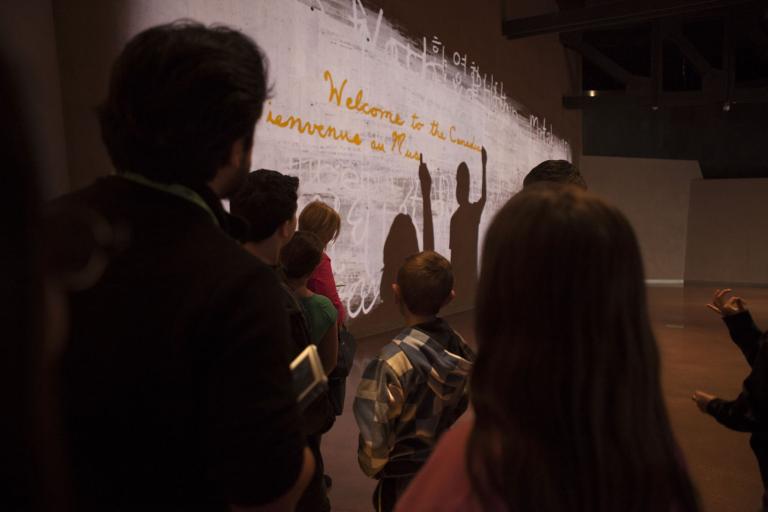A group of people in front of a wall watching a projection. The projection shows two shadowy silhouettes in the process of writing the phrase “Welcome to the Canadian Museum for Human Rights” in English and French. Partially obscured.