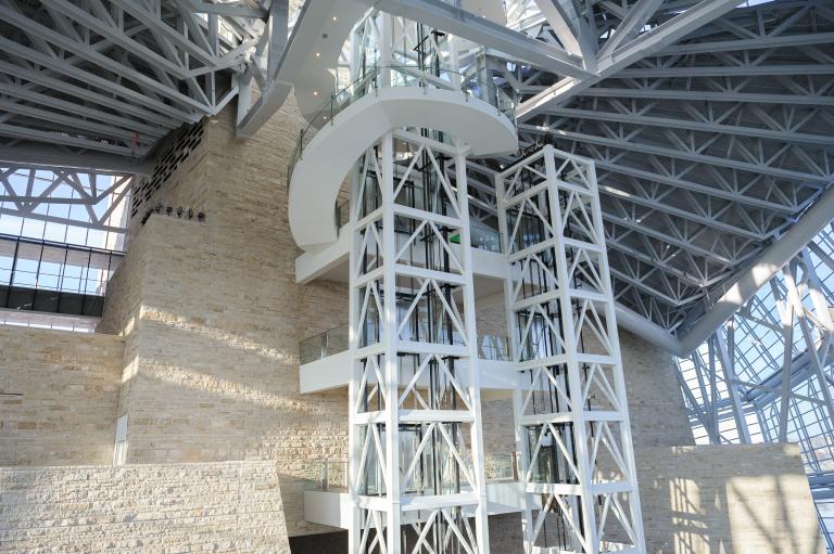 The interior of a large open spaced part of the Museum with two elevator shafts in the centre of the image, and a limestone wall.