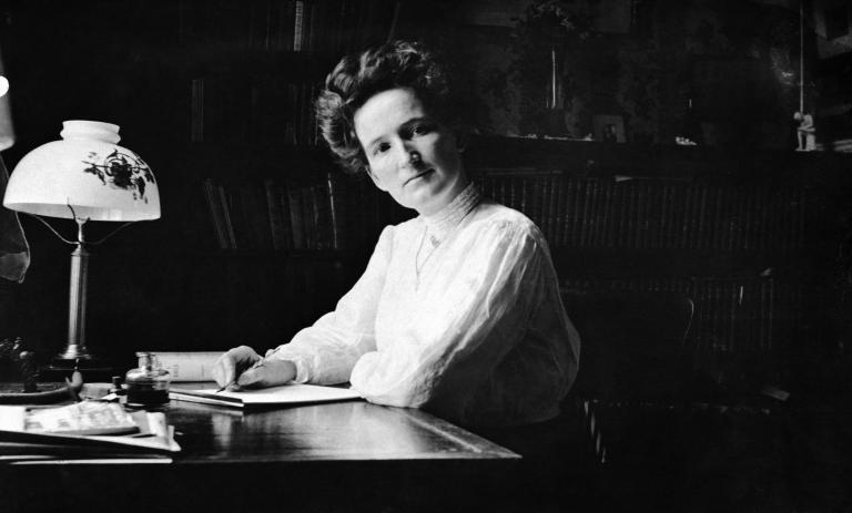 A black and white image of Nellie McClung sitting at a desk. She is wearing a white blouse and holds a pen in her hand. On the desk there is a lamp, an inkwell and some paper. Partially obscured.