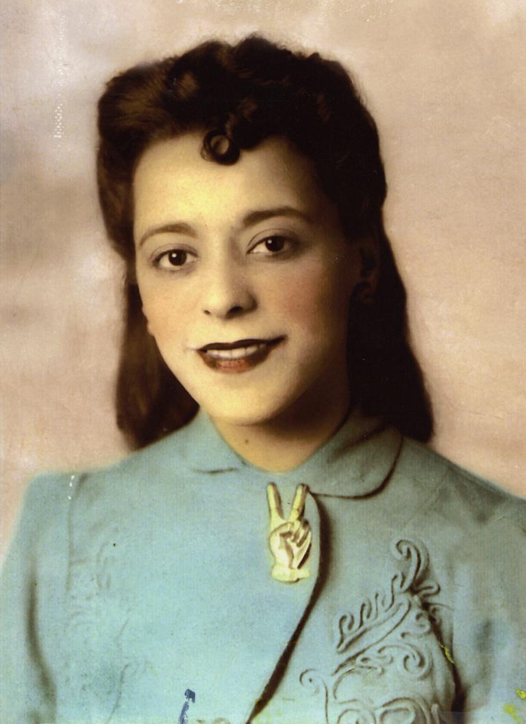 A head-and-shoulder portrait of a smiling Viola Desmond. She is wearing a light blue jacket with embroidered patterns on it. The jacket is held together at the collar by a large pin in the shape of a hand making a “V for victory” symbol. Partially obscured.