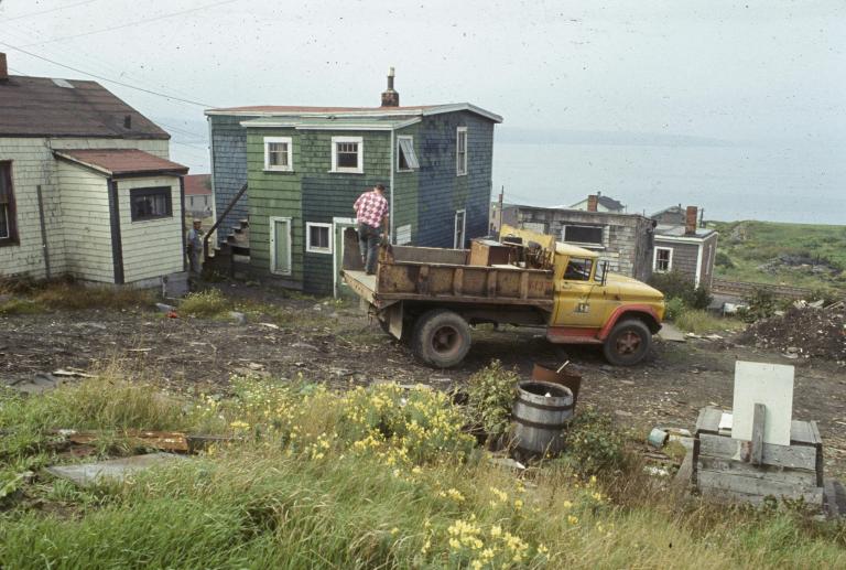 A man stands on the back of a garbage truck that is parked in front of a wooden house with green shingling. In the background rail lines and more houses can be seen, and behind that, Halifax harbor.