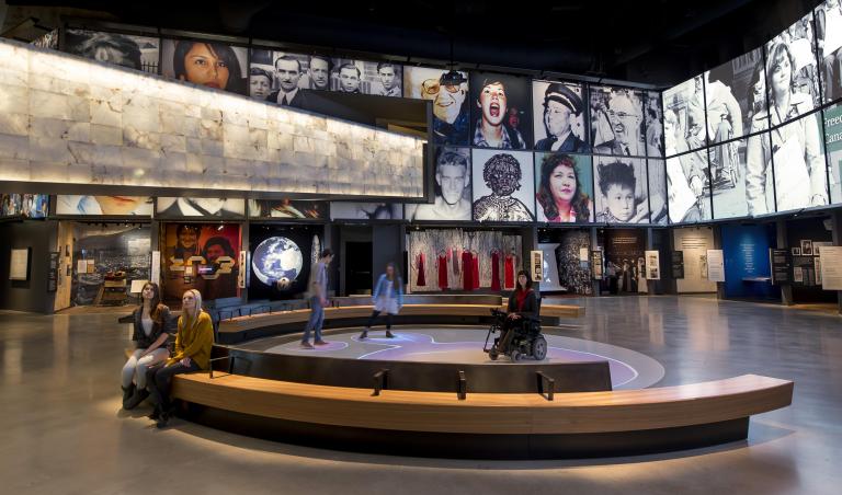 Five people in a museum gallery. Two sit on a bench. Three people, including one in a wheelchair, are on a circular area where bubbles of coloured light are projected on the floor. Partially obscured.