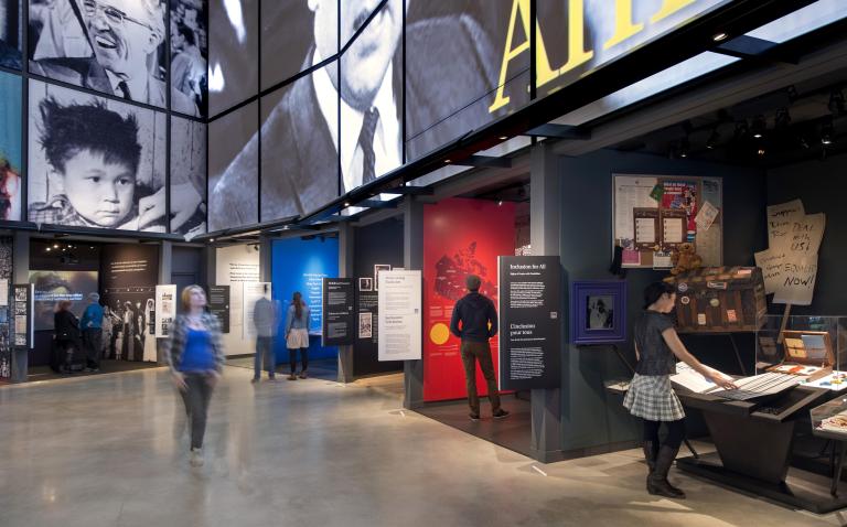 One person is walking in a Museum gallery, while six others are standing in or near alcoves that line the wall of the gallery. Each alcove contains a different exhibit with text or images. Partially obscured.