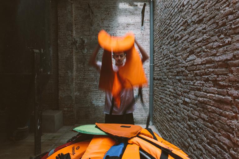 A teenage boy holds a life jacket in his hands while he stands behind a dome-like structure made of lifejackets. Partially obscured.