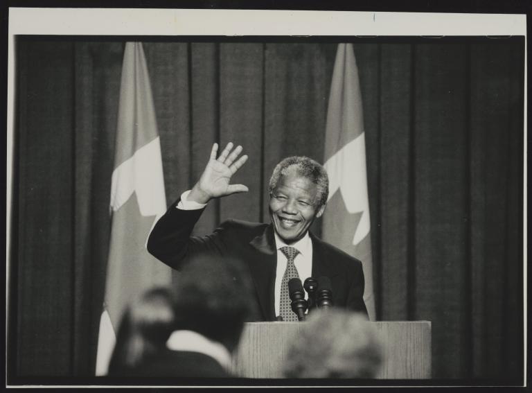 A black and white photograph of Nelson Mandela standing in front of a podium. He is smiling and waving. Partially obscured.