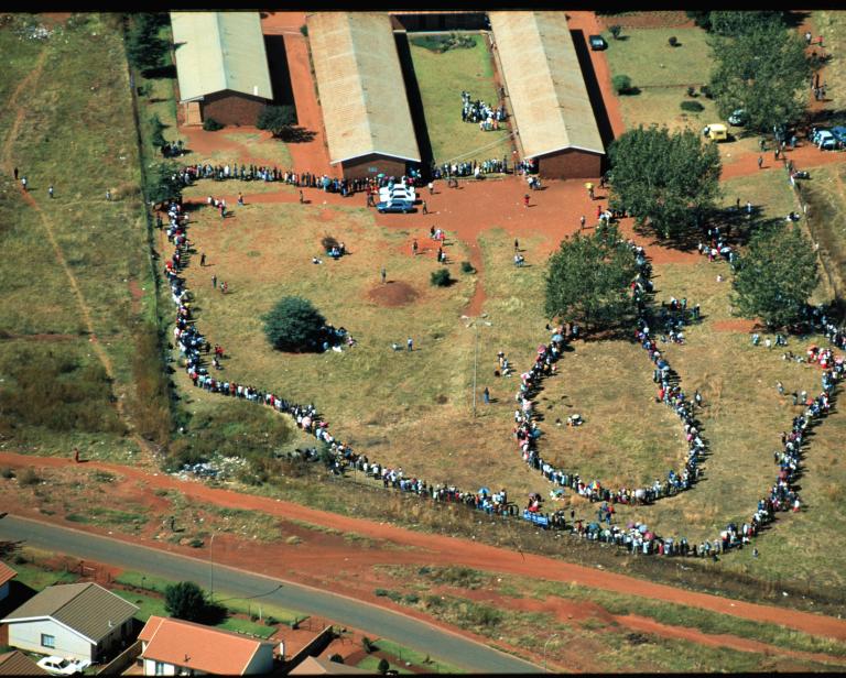 View from above of people standing outside in lines. The ground is mostly covered in grass and the exposed earth is of a reddish colour.