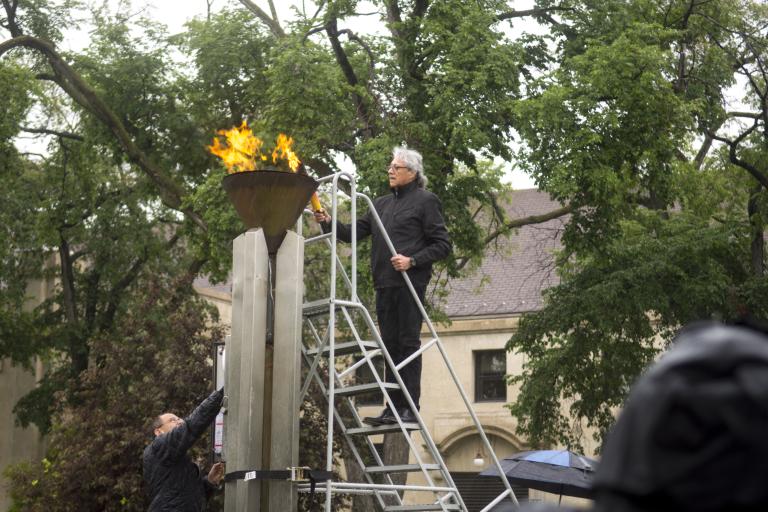 A man carrying a torch stands on a ladder to light a flame within a copper cauldron. Partially obscured.