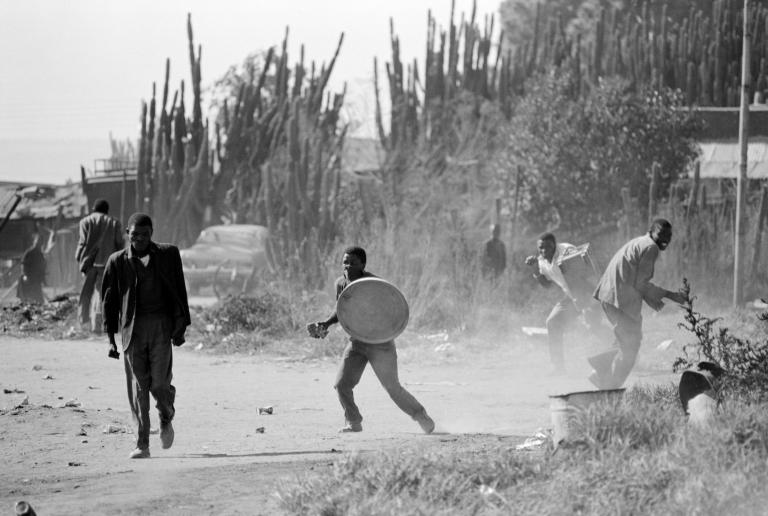 Black and white photo of a few Black men in a dusty outside environment. One is holding up a trashcan lid as a shield.