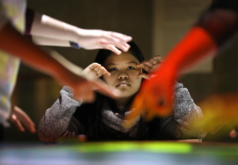 A girl looks forward, resting her elbows on an interactive table with her hands raised to her cheeks. In front of her are several hands reaching towards and pointing to the table. Partially obscured.