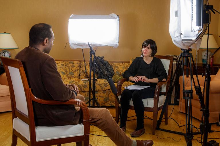 A man and a woman sit in chairs facing each other. There is a microphone on a stand positioned between them. There are also large lights on both sides of the woman’s chair, which are pointed towards the man. Partially obscured.