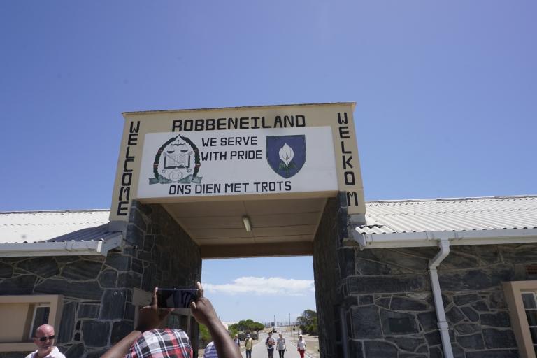 A opening through a building with a large sign over it. The sign reads “Welcome” and “We serve with pride” in both English and Afrikaans. At the top of the sign it says “Robbeneiland.”