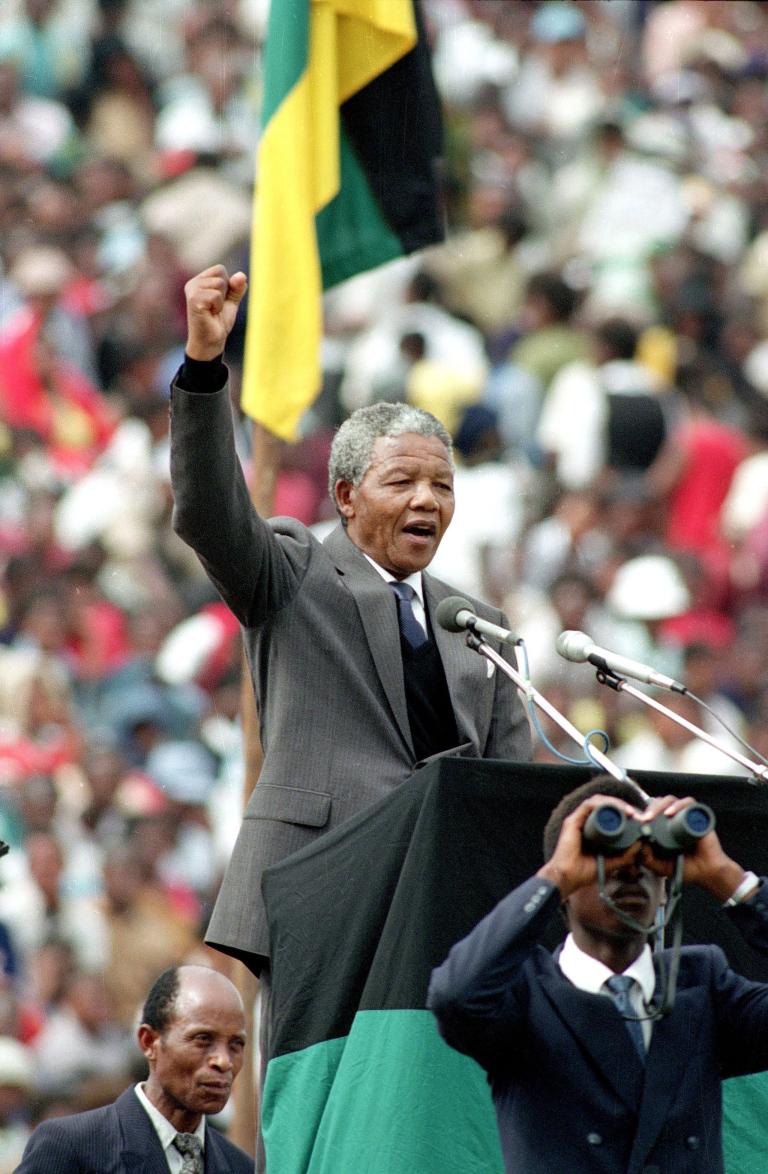 Nelson Mandela, an older dark-skinned man with short grey hair, stands in front of a podium and speaks into two microphones. He is wearing a grey suit with a tie. He is raising his right arm above his head and his hand is making a fist. Partially obscured.