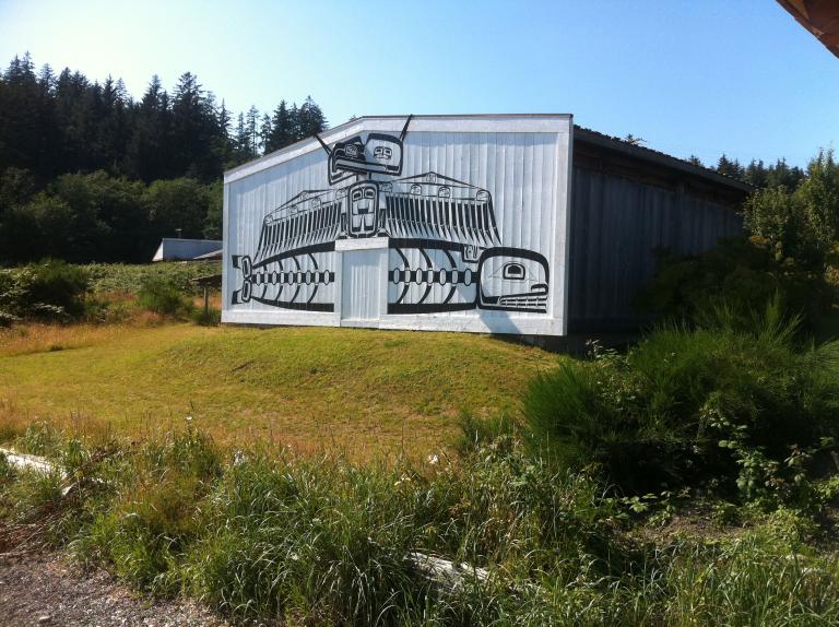 A large rectangular white building sits on a slight rise. The front is painted with stylized representations of a bird sitting on an aquatic creature. The beak of the bird is three-dimensional, coming out of the building’s wall. There is a single white door in the middle of the front, bisecting the aquatic creature and sitting directly below the bird’s feet.