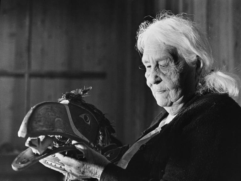 A black and white image of an elderly woman with white hair holding a mask that is shaped and painted to represent a bird’s head. It features a long beak with an open mouth and feathers attached to the top of the head. The woman is looking directly at the mask she is holding. Partially obscured.