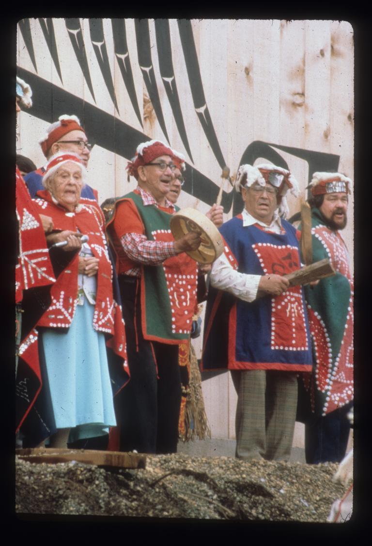 A group of older men and one woman sing while standing in front of a large wooden building. One man is playing a drum. The singers are wearing long shirts or blankets that feature patterns made from buttons.