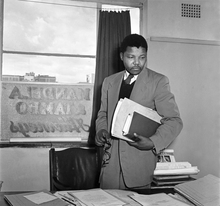 A-black-and-white image of Nelson Mandela, a young black man wearing a suit and carrying a book and some files under his left arm, standing behind a desk in an office.