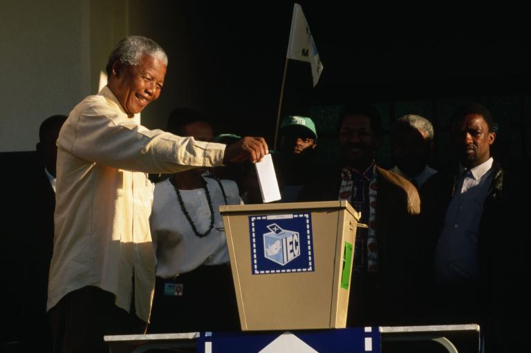 An elderly Nelson Mandela smiles as he reaches out with his right arm to put a ballot in a large metal ballot box.