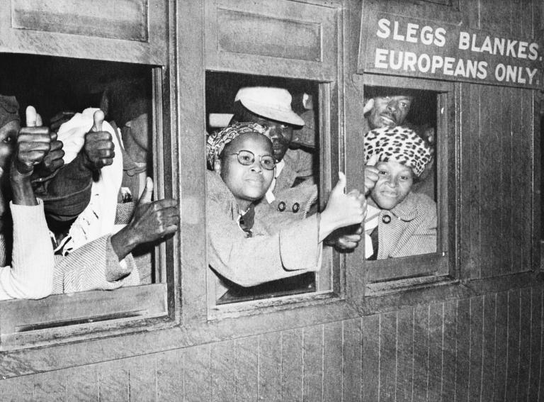 A black-and-white image of a group of black men and women look out a set of train car windows. Many are holding out their hands and making a “thumbs up” sign. A sign above the train window reads “Europeans only.”
