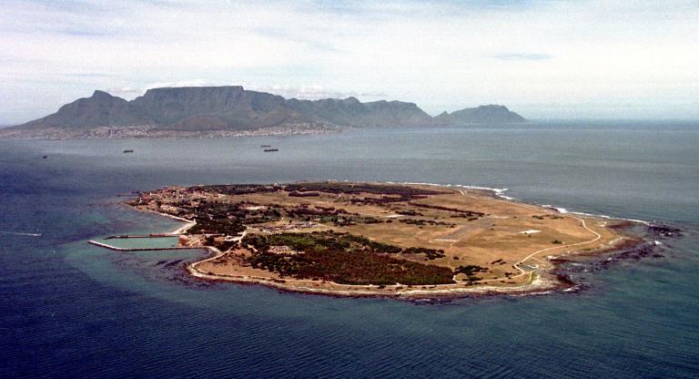 An aerial view of Robben Island. The island is very flat and roughly oval in shape, with a little under half its surface covered with trees. An airfield is visible on the right side of the island. In the distant background behind the island, the mainland can be seen, including the buildings of a city. Rising above the city are large mountains. 