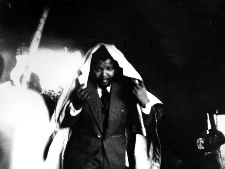 A black-and-white image of a young Nelson Mandela wearing a suit, crouching and holding a trench coat around his head, as if he is hiding himself from rain.