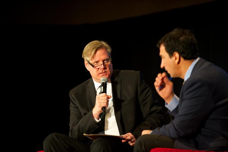 Two men sitting in front of a black backdrop. One man is speaking into a microphone. Partially obscured.
