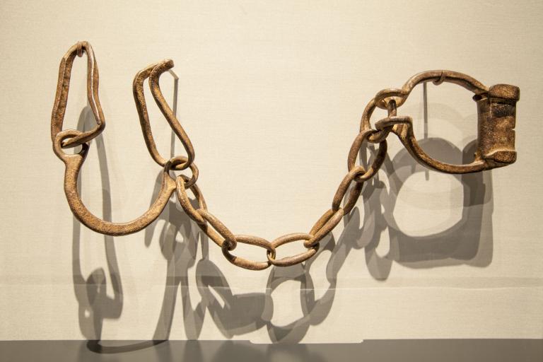 Rusted iron manacles hang on hooks on a plain white wall. Partially obscured.